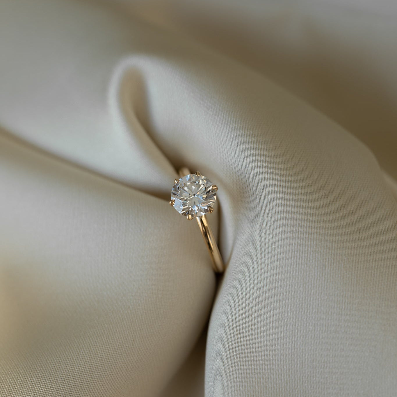Gold solitaire ring with a round diamond resting elegantly on draped ivory fabric.