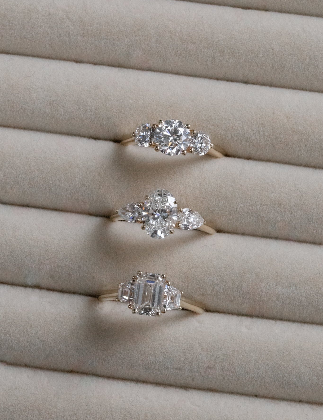 Three gold engagement rings with different diamond cuts (round, pear, emerald) displayed in a light velvet ring holder.