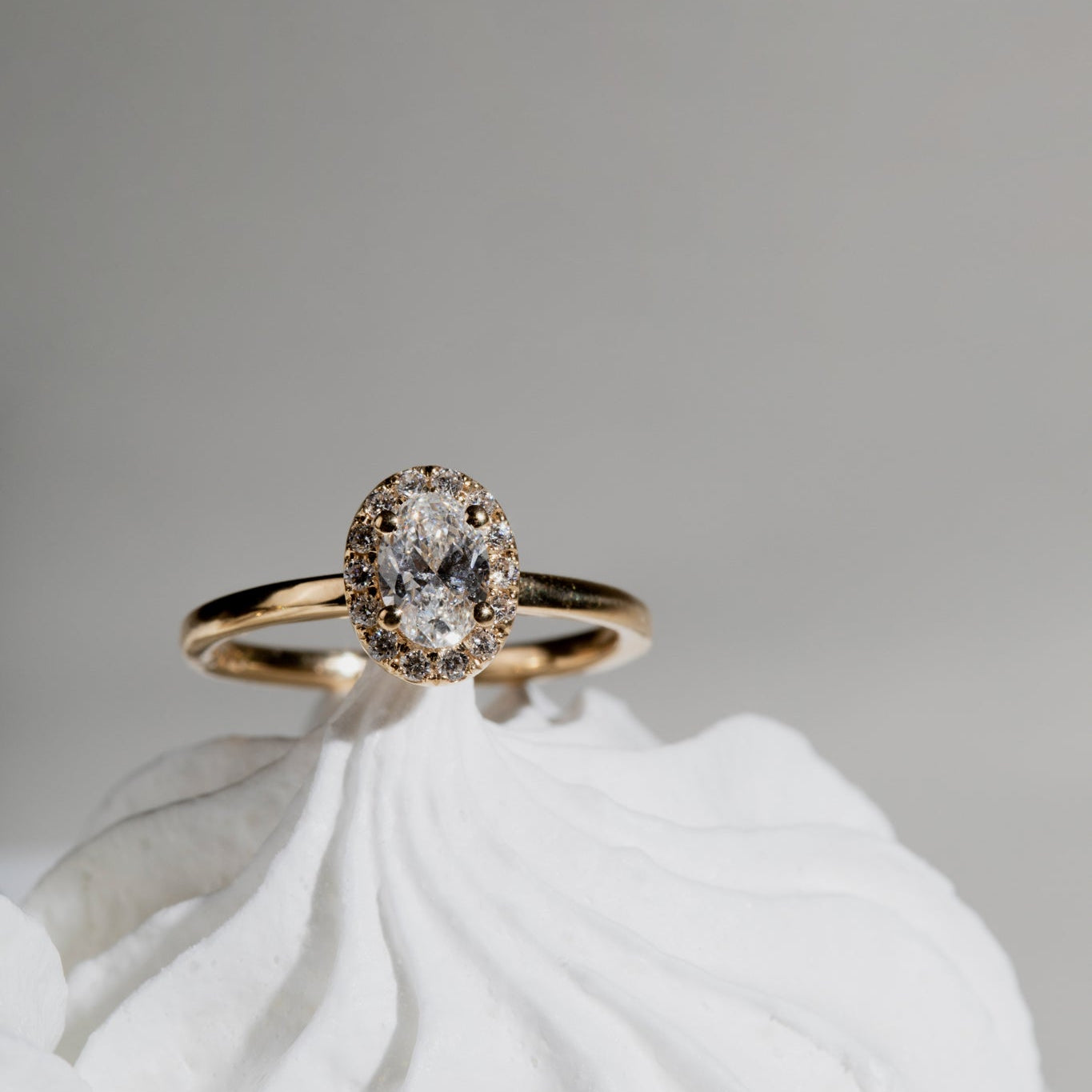 Gold ring with an oval diamond centerpiece surrounded by a halo of smaller diamonds, displayed on a white textured background.