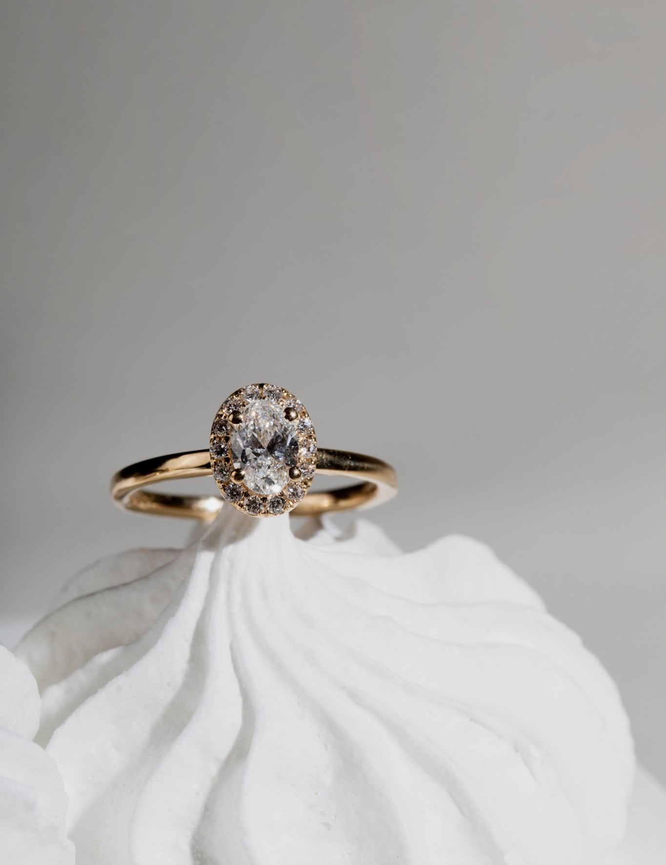 Gold ring with an oval diamond centerpiece surrounded by a halo of smaller diamonds, displayed on a white textured background.