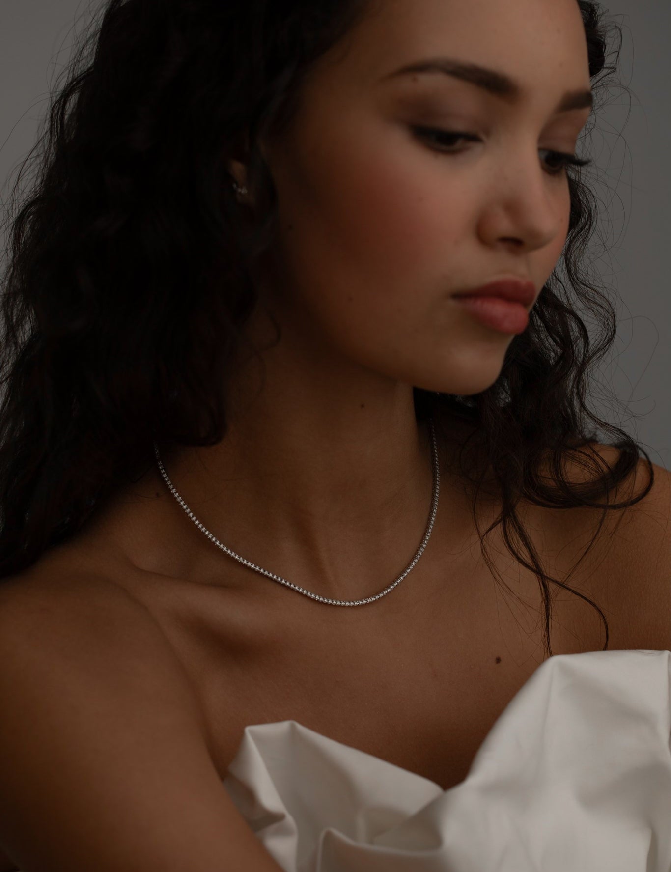 Woman with wavy dark hair wearing a delicate diamond tennis necklace, styled with a soft white fabric.