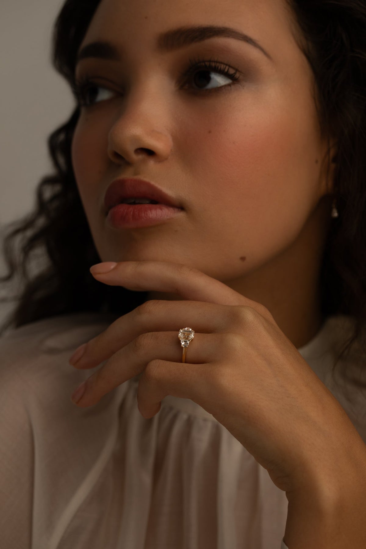 Woman wearing a gold ring with a pear-shaped diamond, gazing thoughtfully, dressed in a soft white blouse.