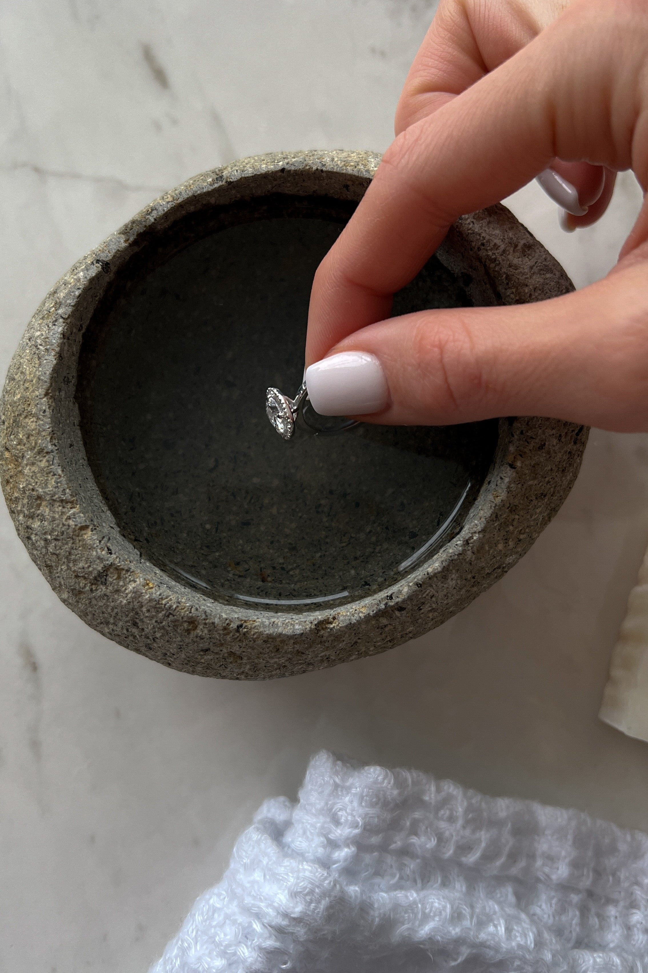 A hand with white-painted nails dipping a silver ring into a small round stone bowl filled with water, placed on a marble surface.