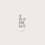 Silver pendant in the shape of the letter "K," adorned with sparkling gemstones, displayed on a light beige background.