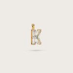 Gold pendant in the shape of the letter "K," adorned with sparkling gemstones, displayed on a light beige background.
