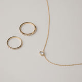 A minimalist flat-lay product photograph featuring a round bezel-set diamond pendant necklace displayed beside two matching delicate gold pavé diamond stacking rings on a light grey background.