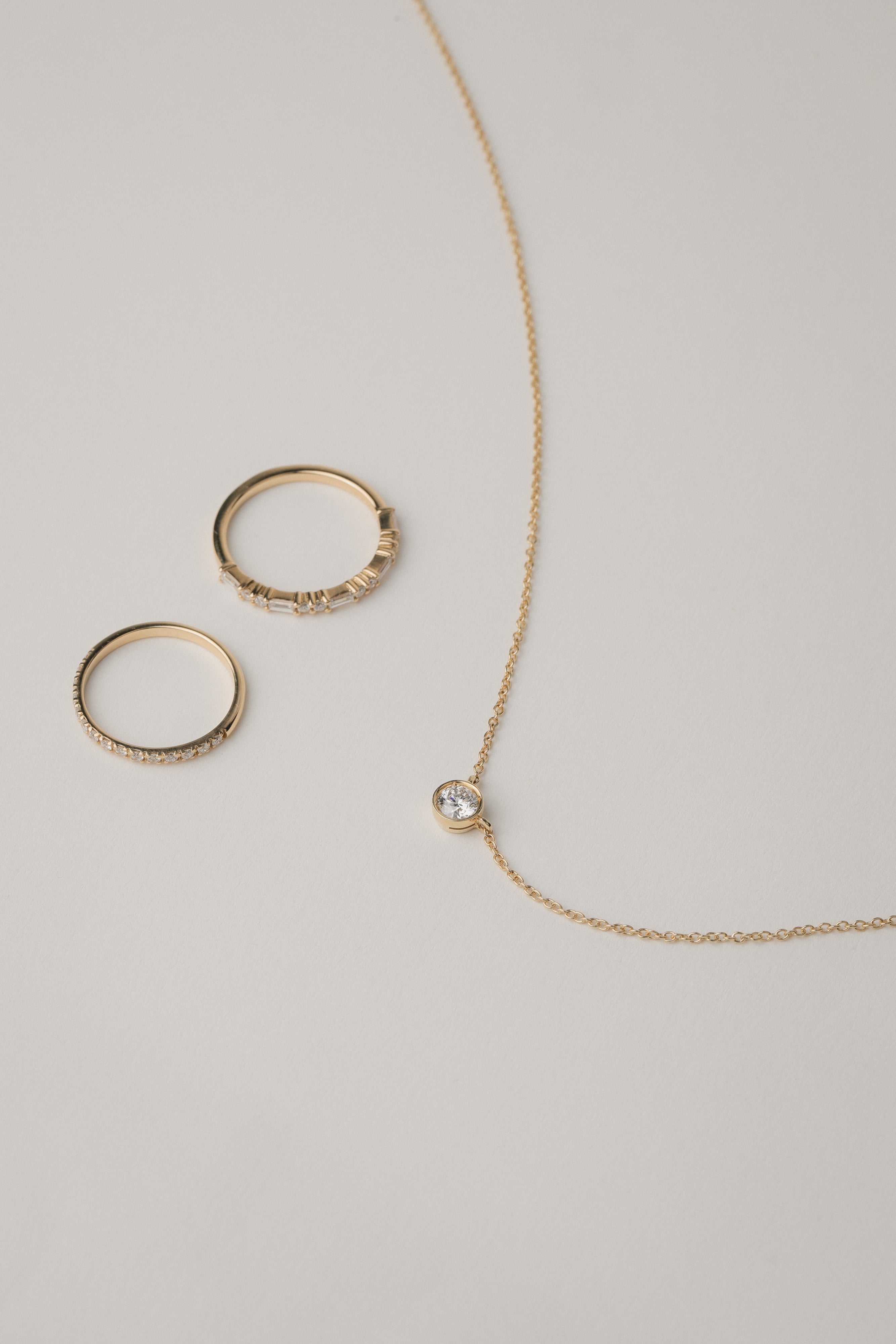 A minimalist flat-lay product photograph featuring a round bezel-set diamond pendant necklace displayed beside two matching delicate gold pavé diamond stacking rings on a light grey background.
