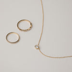 A minimalist flat-lay product photograph featuring a round white gold bezel-set diamond pendant necklace displayed beside two matching delicate gold pavé diamond stacking rings on a light grey background.
