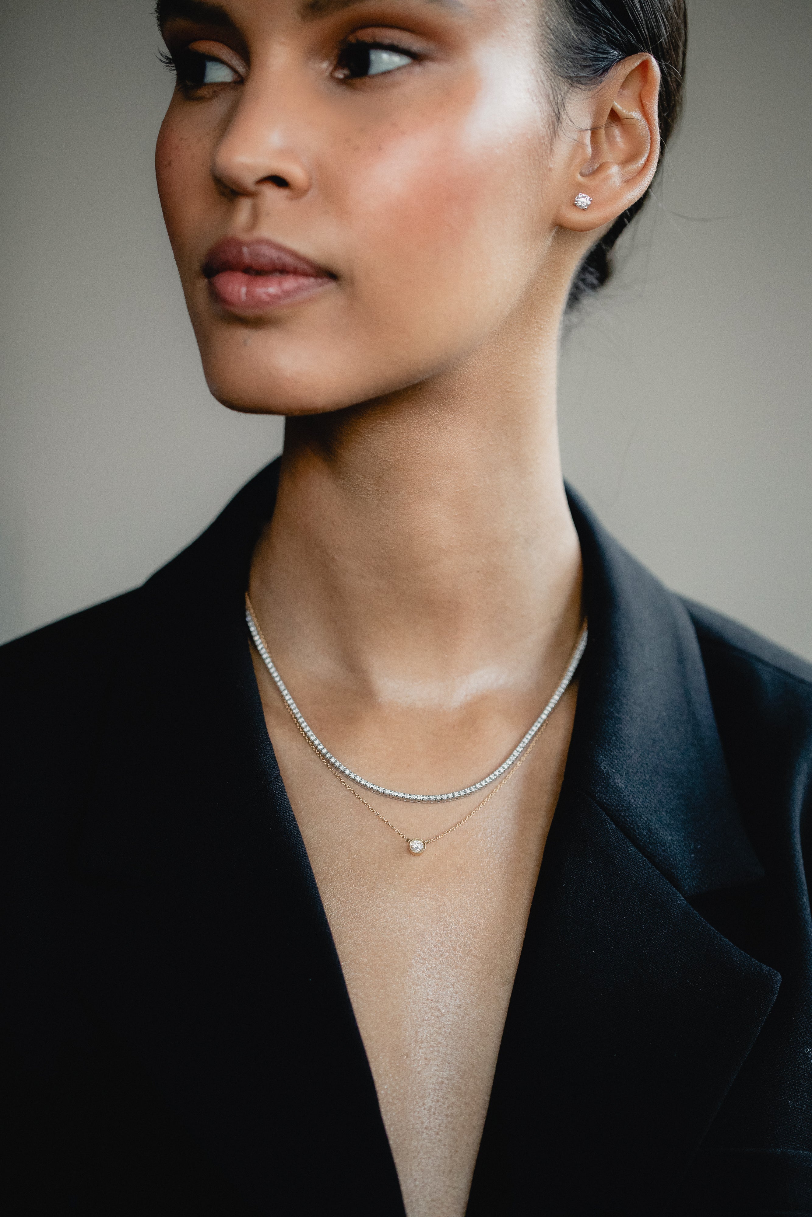 An elegant editorial lifestyle portrait of a model wearing a blazer, showcasing a luxury layering combination with a white gold bezel-set diamond necklace layered beneath a shimmering white gold diamond tennis necklace.