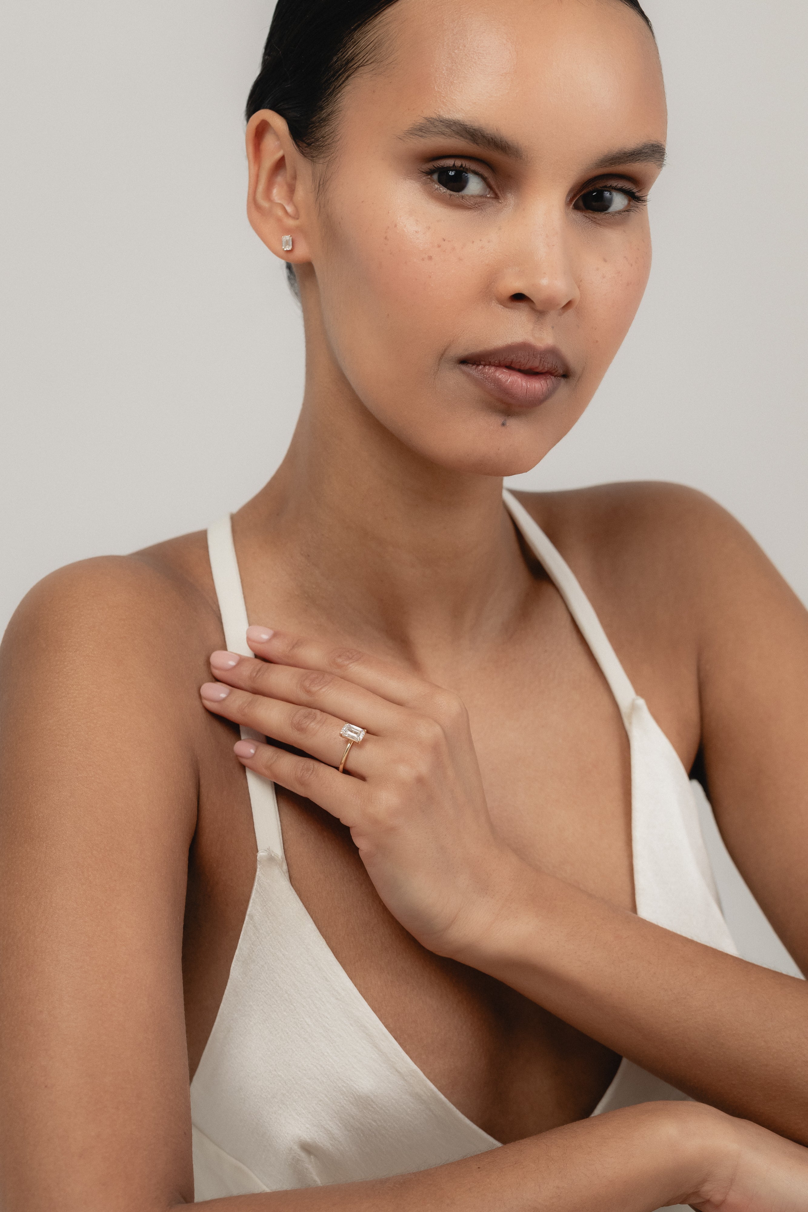 An editorial portrait of a model wearing a white strap top, elegantly displaying a white gold KARAAT emerald-shaped diamond solitaire ring stacked with a gold band on her finger.