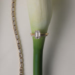 An artistic still-life photograph of a yellow gold KARAAT emerald-cut diamond engagement ring slipped onto the stem of a white calla lily, placed next to a shimmering diamond tennis bracelet.