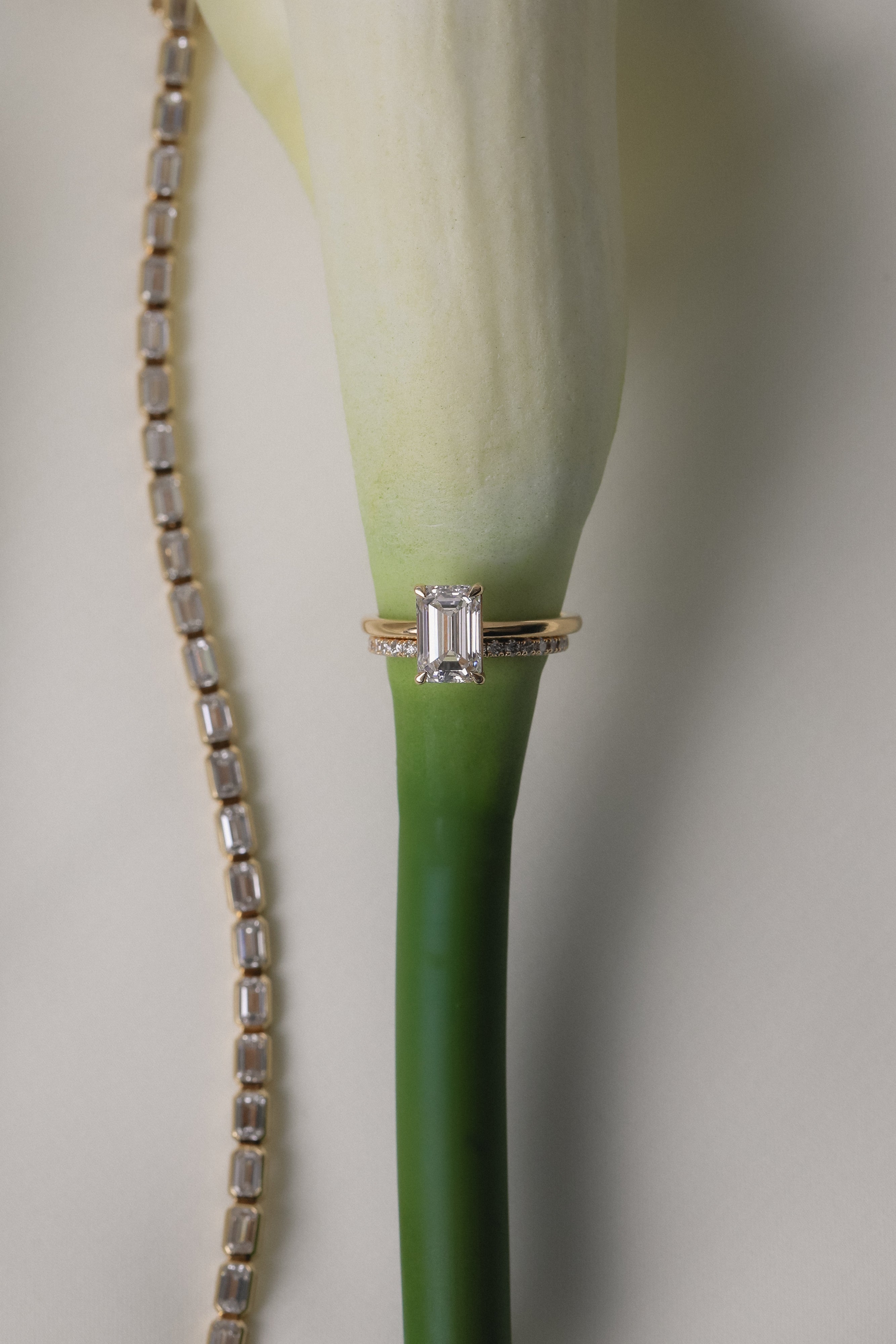 An artistic still-life photograph of a white gold KARAAT emerald-cut diamond engagement ring slipped onto the stem of a white calla lily, placed next to a shimmering diamond tennis bracelet.