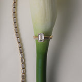 An artistic still-life photograph of a rose gold KARAAT emerald-cut diamond engagement ring slipped onto the stem of a white calla lily, placed next to a shimmering diamond tennis bracelet.