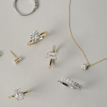 A luxurious flat-lay arrangement of KARAAT fine jewelry on a neutral grey background, featuring a white gold emerald-cut engagement ring, an oval solitaire, a round diamond ring, a tennis bracelet, and a delicate diamond pendant necklace.