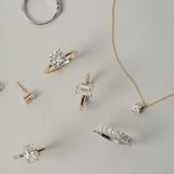 A luxurious flat-lay arrangement of KARAAT fine jewelry on a neutral grey background, featuring a white gold emerald-cut engagement ring, an oval solitaire, a round diamond ring, a tennis bracelet, and a delicate diamond pendant necklace.