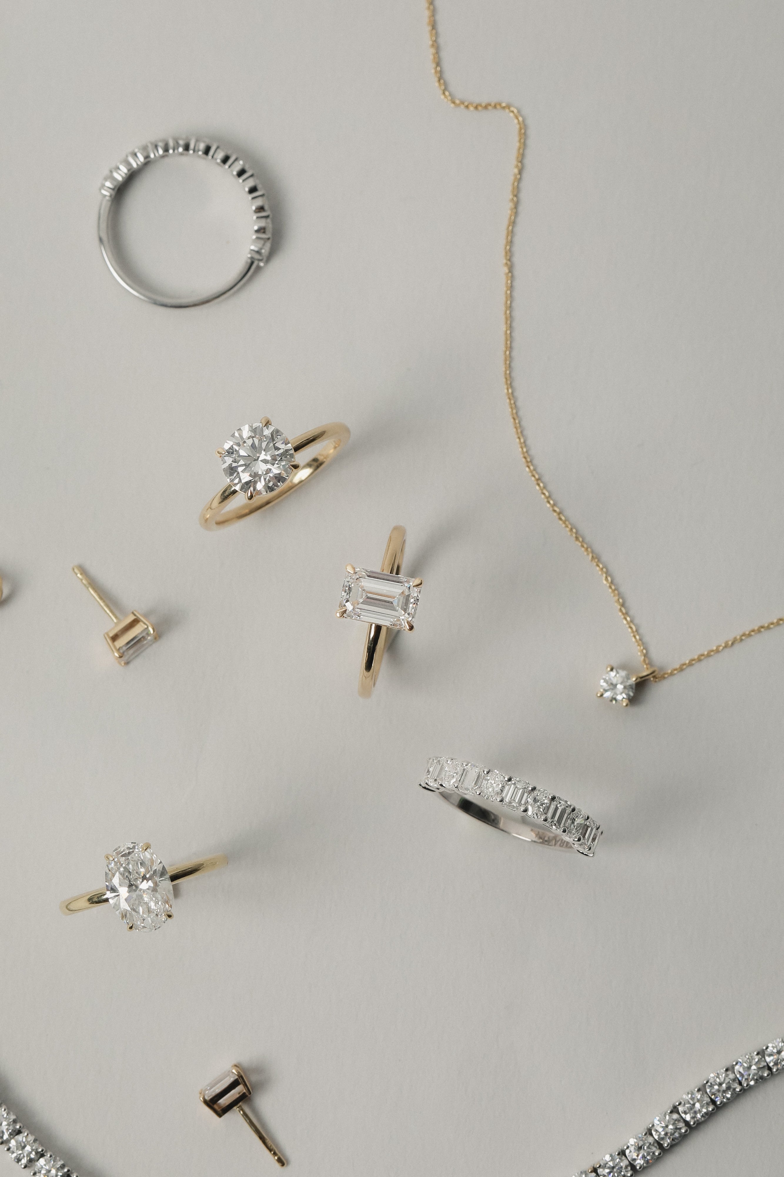 A luxurious flat-lay arrangement of KARAAT fine jewelry on a neutral grey background, featuring a white gold emerald-cut engagement ring, an oval solitaire, a round diamond ring, a tennis bracelet, and a delicate diamond pendant necklace.