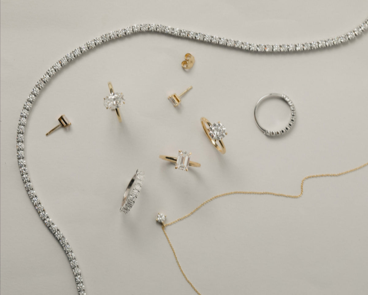A styled flat-lay of multiple jewelry pieces on a light warm-gray background, including a white gold tennis necklace, a yellow gold pendant necklace, several rings, stud earrings, and a small pendant.