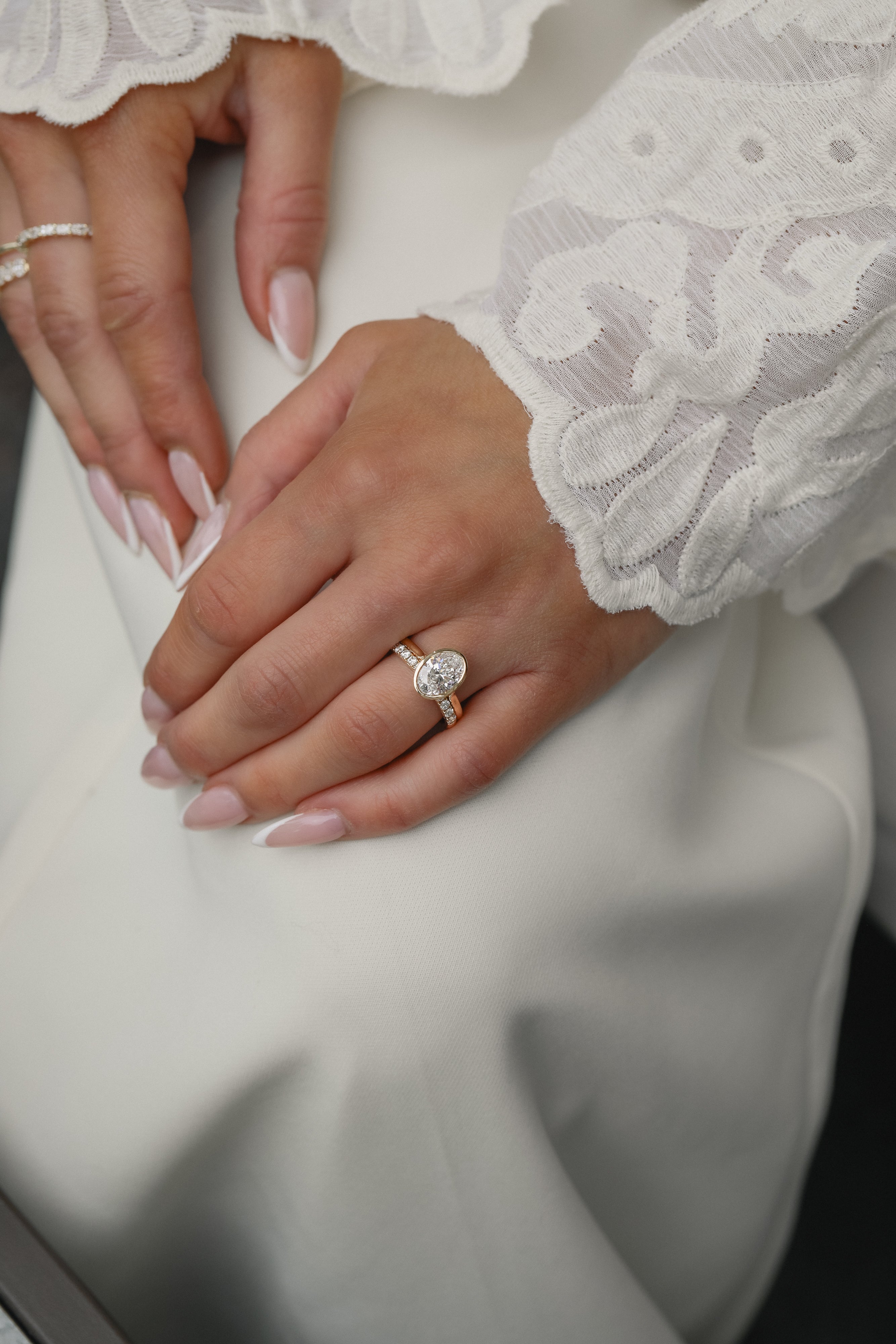 Elegant bridal lifestyle shot of a bride in a lace gown showcasing a modern bezel-set oval diamond engagement ring stacked with the Pavé Diamond Ring II in white gold.