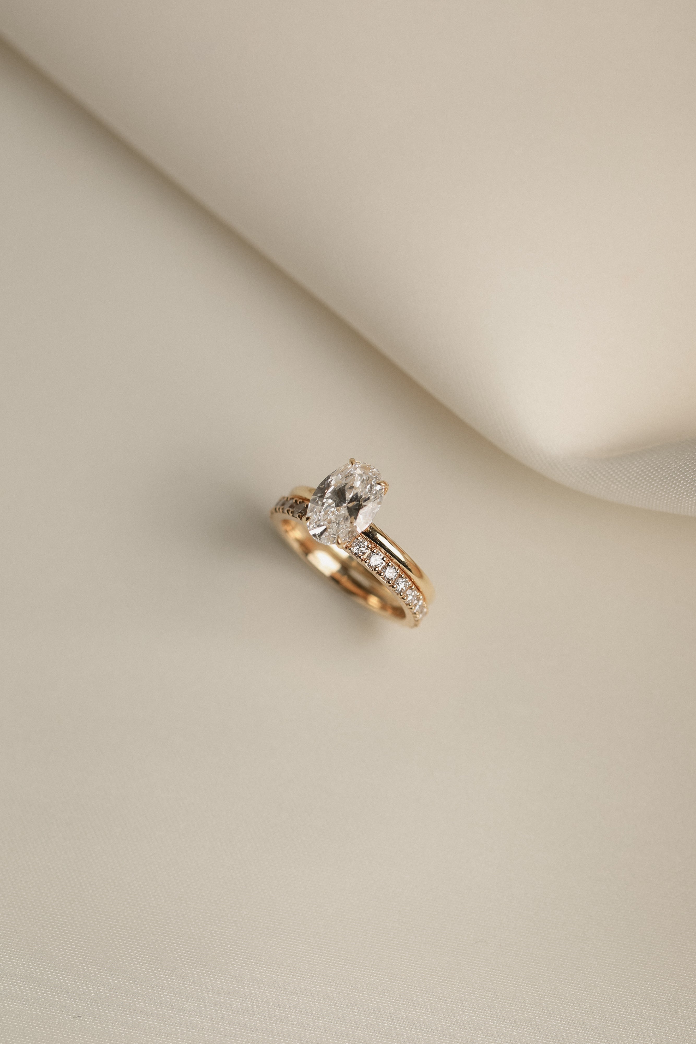 Still life shot of a white gold luxury bridal ring stack featuring a classic oval solitaire diamond engagement ring paired flush with the Pavé Diamond Ring II on a cream fabric background.
