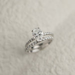 A professional still-life photograph of a KARAAT's oval-cut diamond solitaire engagement ring with a sparkling pavé band in 18k white gold, displayed on a neutral background.