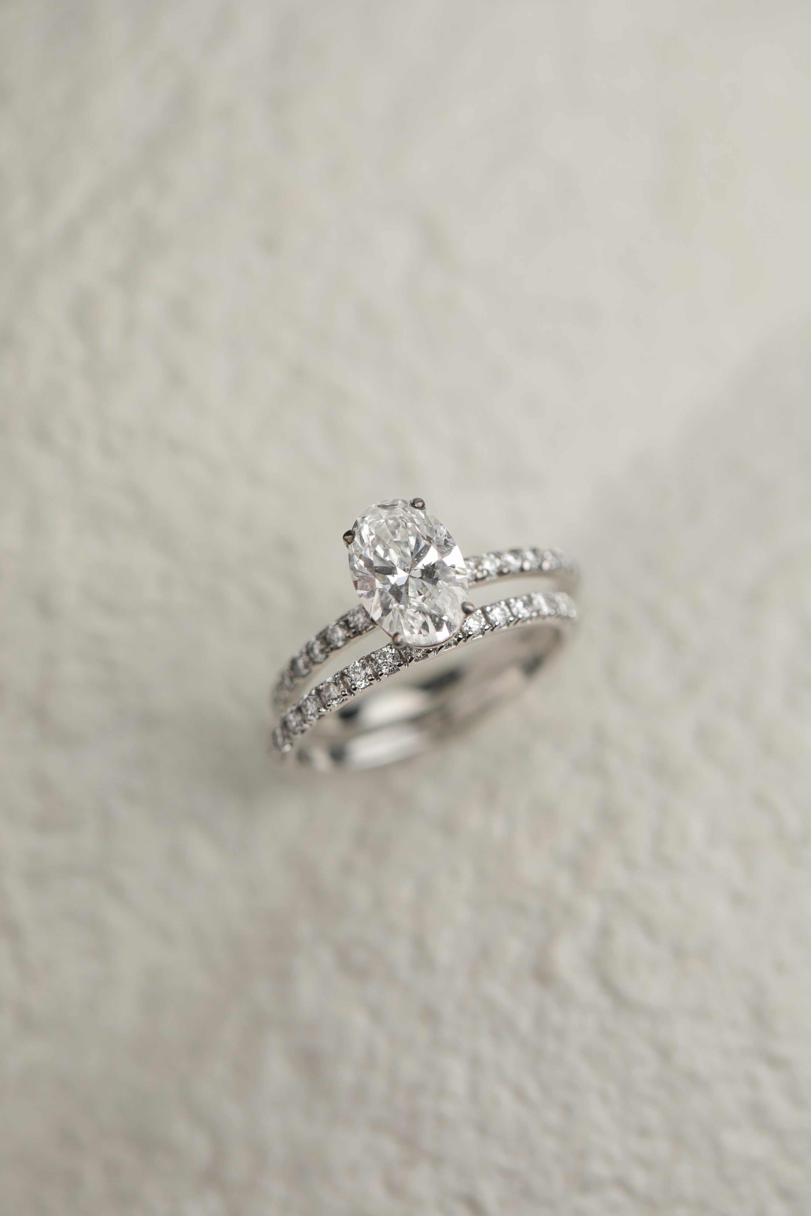 A professional still-life photograph of a KARAAT's oval-cut diamond solitaire engagement ring with a sparkling pavé band in 18k white gold, displayed on a neutral background.