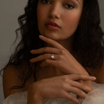 A close-up portrait of a woman with curly hair, wearing a white off-the-shoulder dress. She is gently touching her face with one hand, and a sparkling diamond ring is visible on her finger.