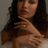 A close-up portrait of a woman with curly hair, wearing a white off-the-shoulder dress. She is gently touching her face with one hand, and a sparkling diamond ring is visible on her finger.