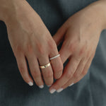 Detail shot of a model's hands gently clasped over a slate blue dress, showing a modern mixed stack featuring a wide solid gold band paired with the striking Pavé Diamond Ring III in yellow gold.