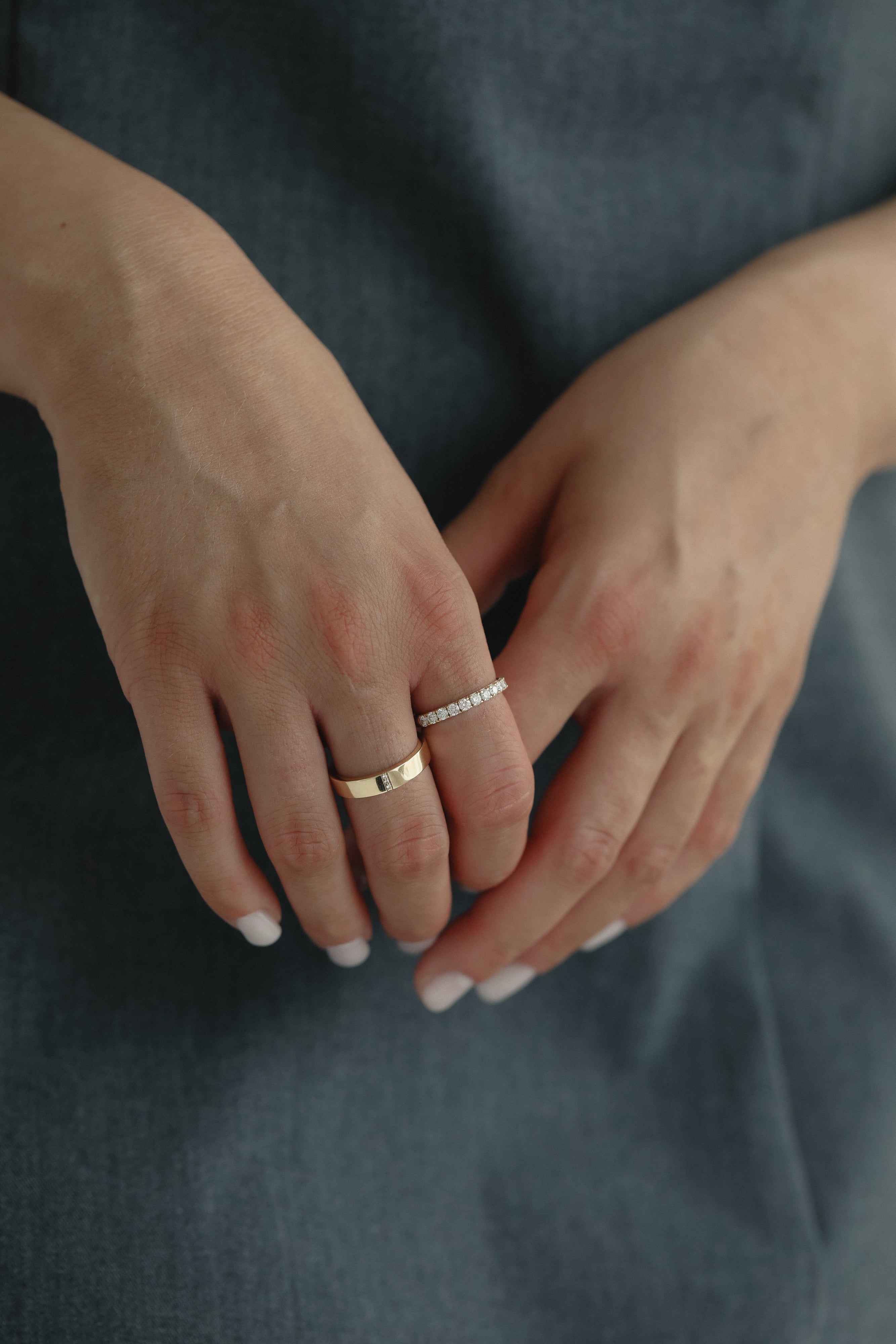 Detail shot of a model's hands gently clasped over a slate blue dress, showing a modern mixed stack featuring a wide solid gold band paired with the striking Pavé Diamond Ring III in yellow gold.