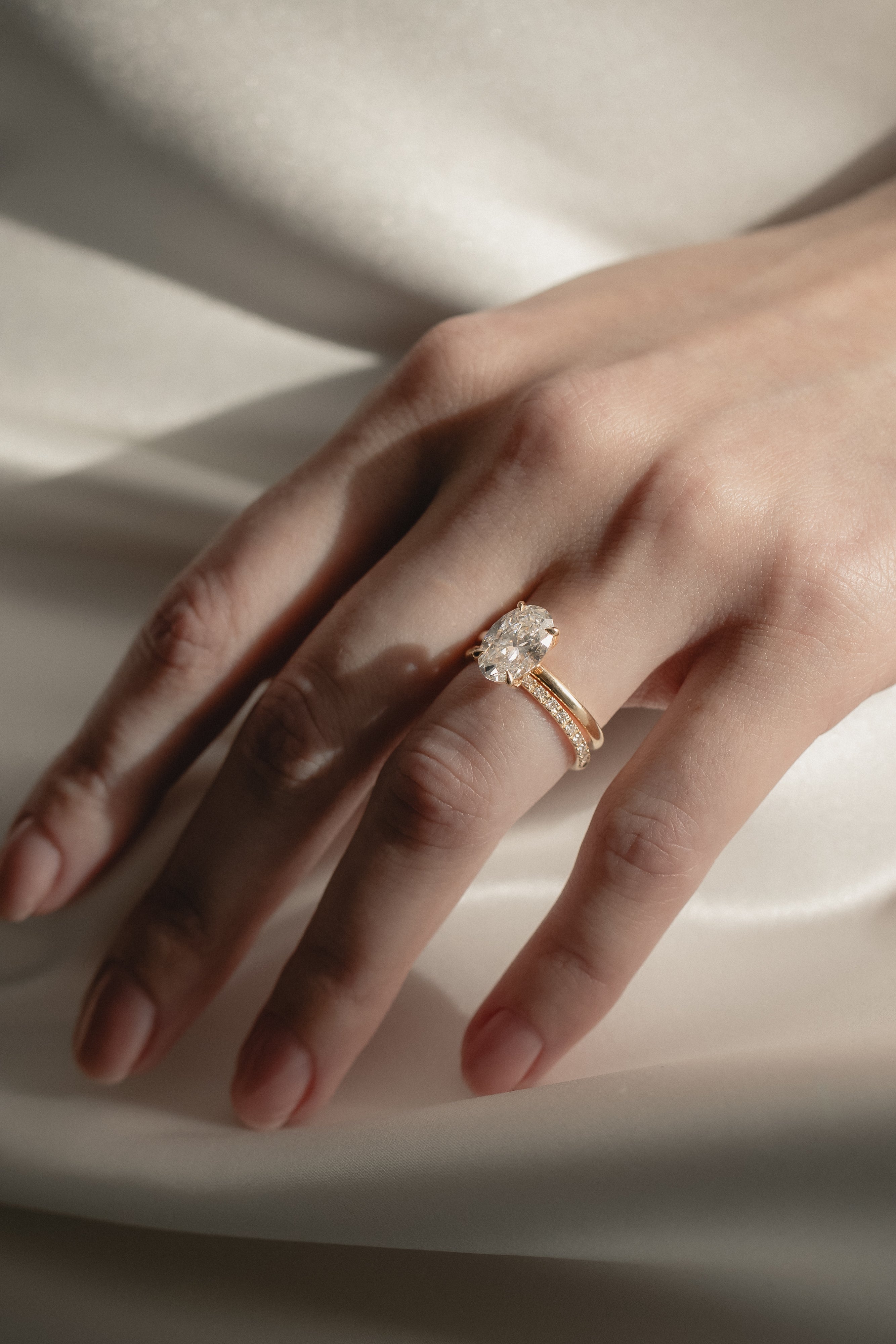 A soft-focus bridal image of a model's hand on shimmering white silk, showcasing a classic rose gold oval solitaire diamond engagement ring nested flush beside a delicate KARAAT pavé diamond wedding band.