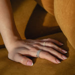 Sunlit detail shot of a model's hand resting on golden ochre fabric, showcasing a prominent ring stack featuring a thick diamond eternity band layered with a delicate classic gold pavé diamond ring.