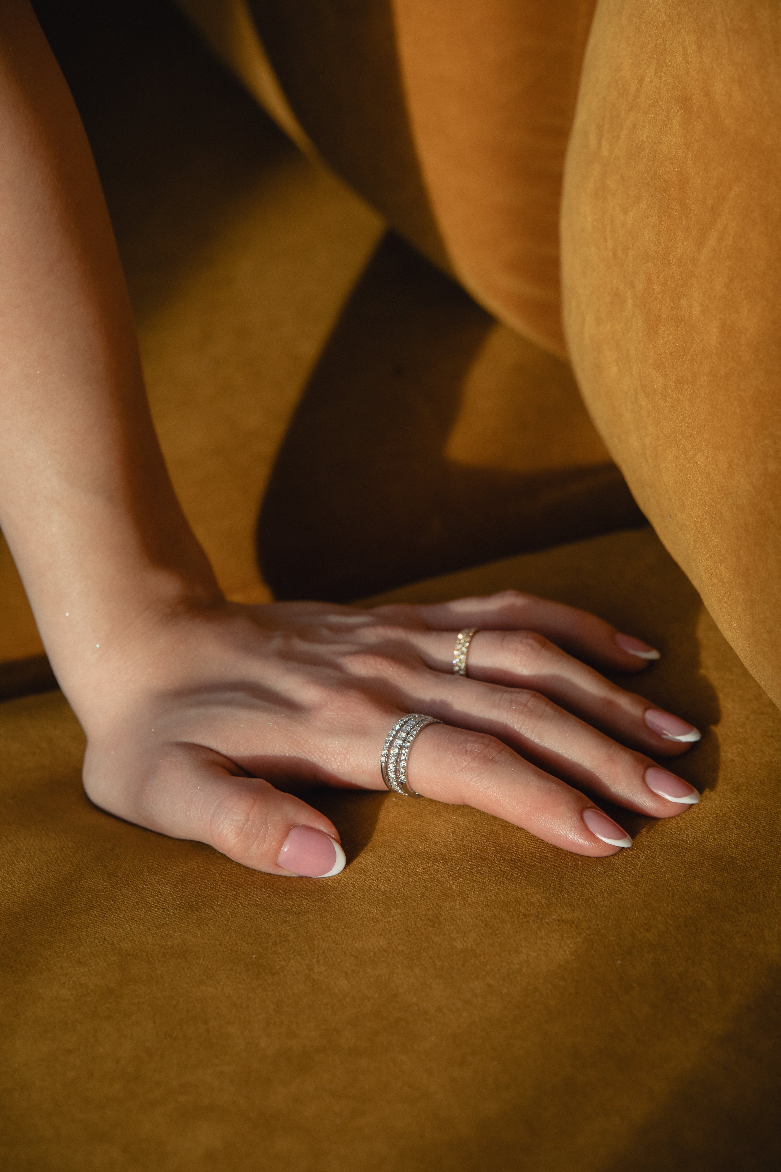 Sunlit detail shot of a model's hand resting on golden ochre fabric, showcasing a prominent ring stack featuring a thick diamond eternity band layered with a delicate classic rose gold pavé diamond ring.