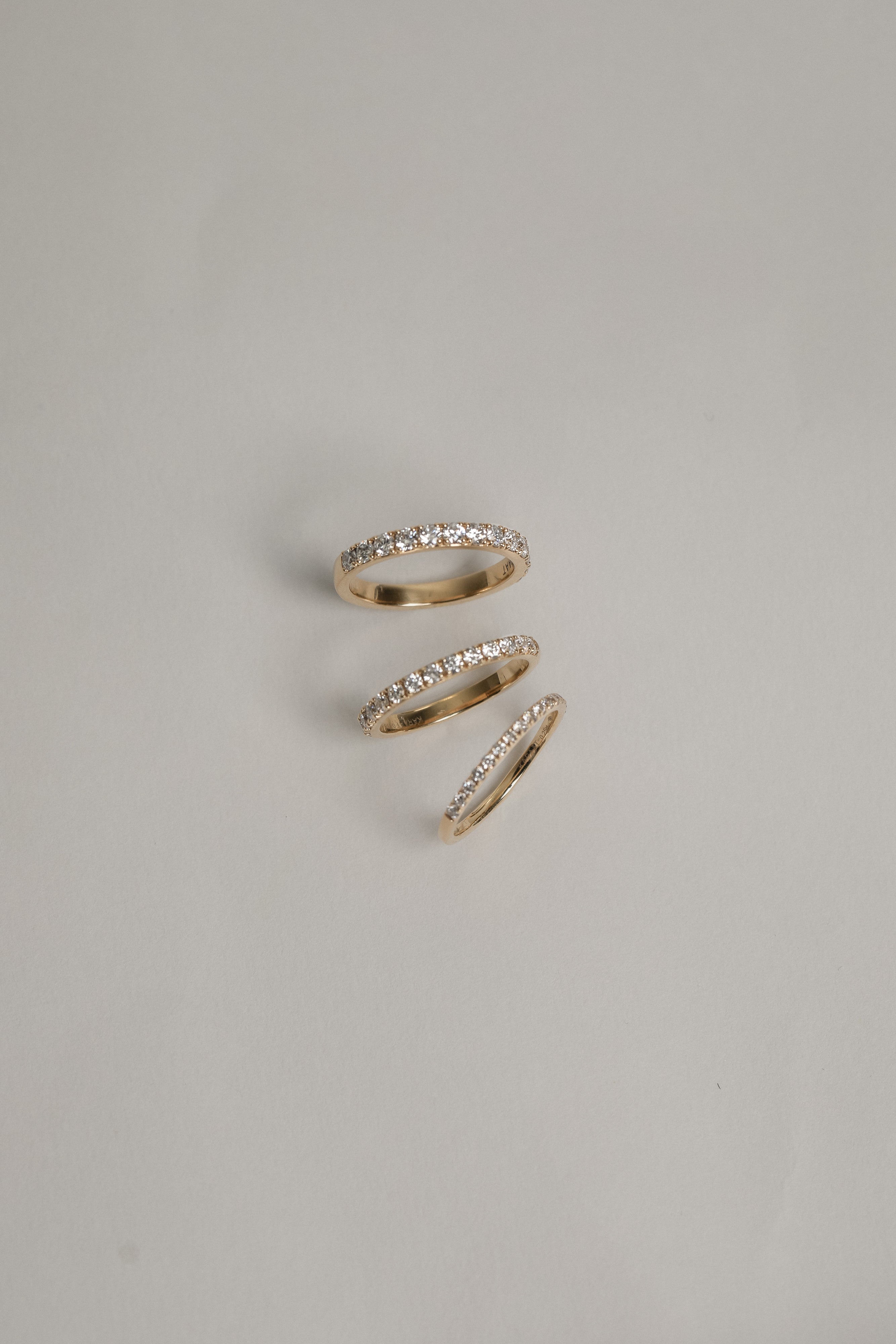 A clean flat-lay product shot displaying three KARAAT pavé diamond rings on a light grey background, showcasing different band widths and diamond carat sizes in polished white gold.