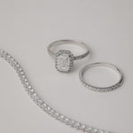 A luxury still-life photograph featuring an emerald-cut diamond halo engagement ring paired with a matching KARAAT white gold pavé diamond ring and a partial tennis bracelet on a neutral grey background.