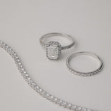A luxury still-life photograph featuring an emerald-cut diamond halo engagement ring paired with a matching KARAAT white gold pavé diamond ring and a partial tennis bracelet on a neutral grey background.