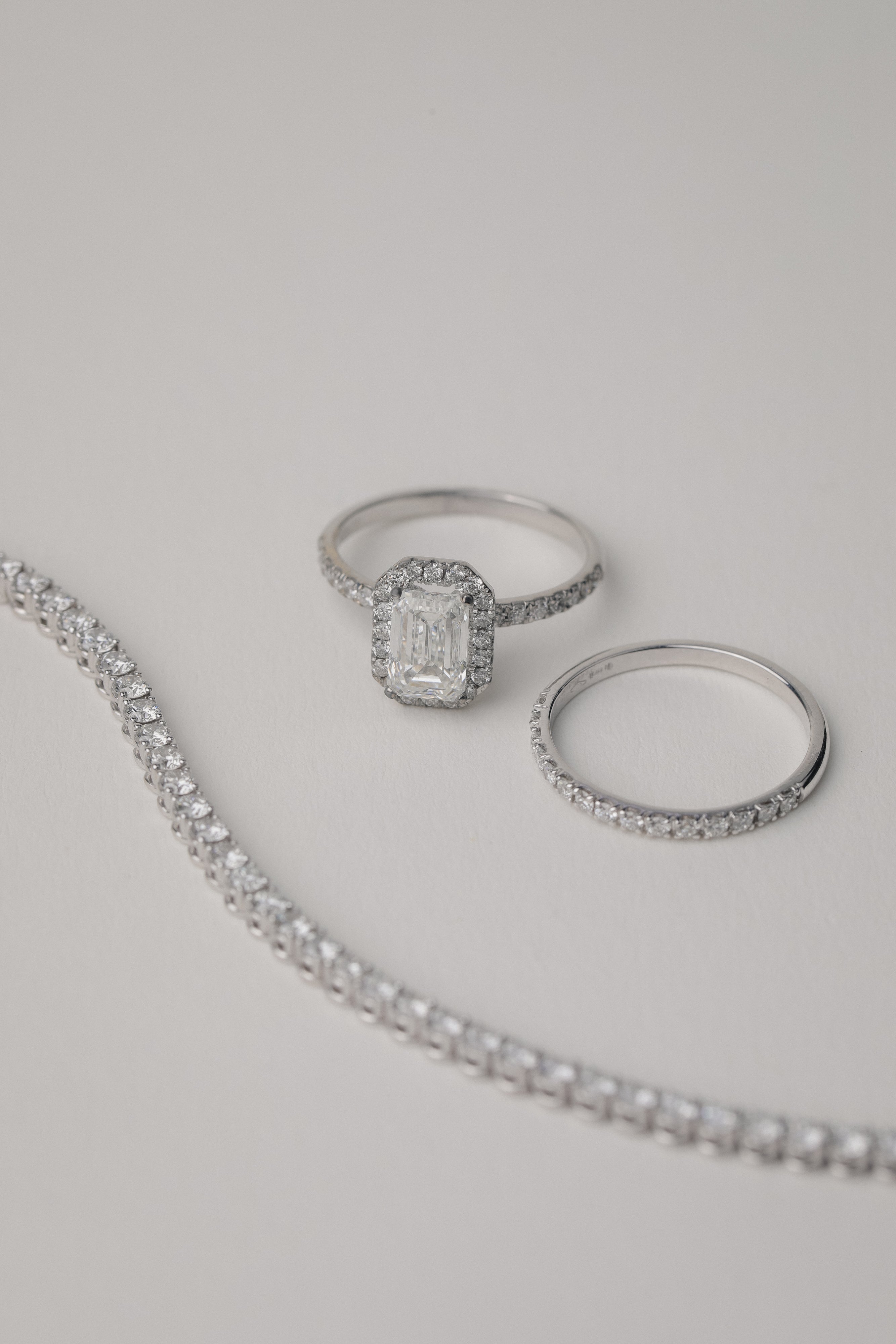 A luxury still-life photograph featuring an emerald-cut diamond halo engagement ring paired with a matching KARAAT white gold pavé diamond ring and a partial tennis bracelet on a neutral grey background.