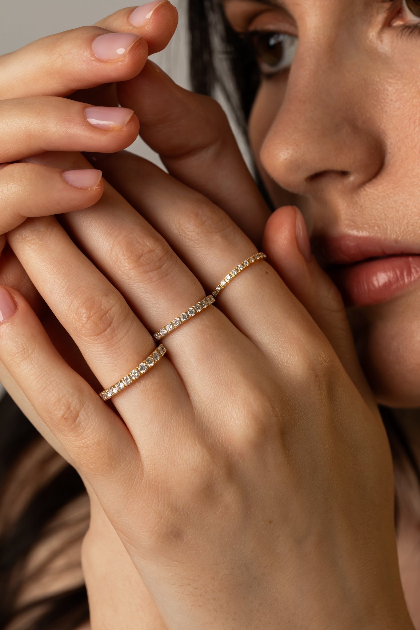 A close-up lifestyle shot of a model holding her hands up near her face, showcasing three delicate KARAAT pavé diamond rings stacked on different fingers in 18k rose gold.