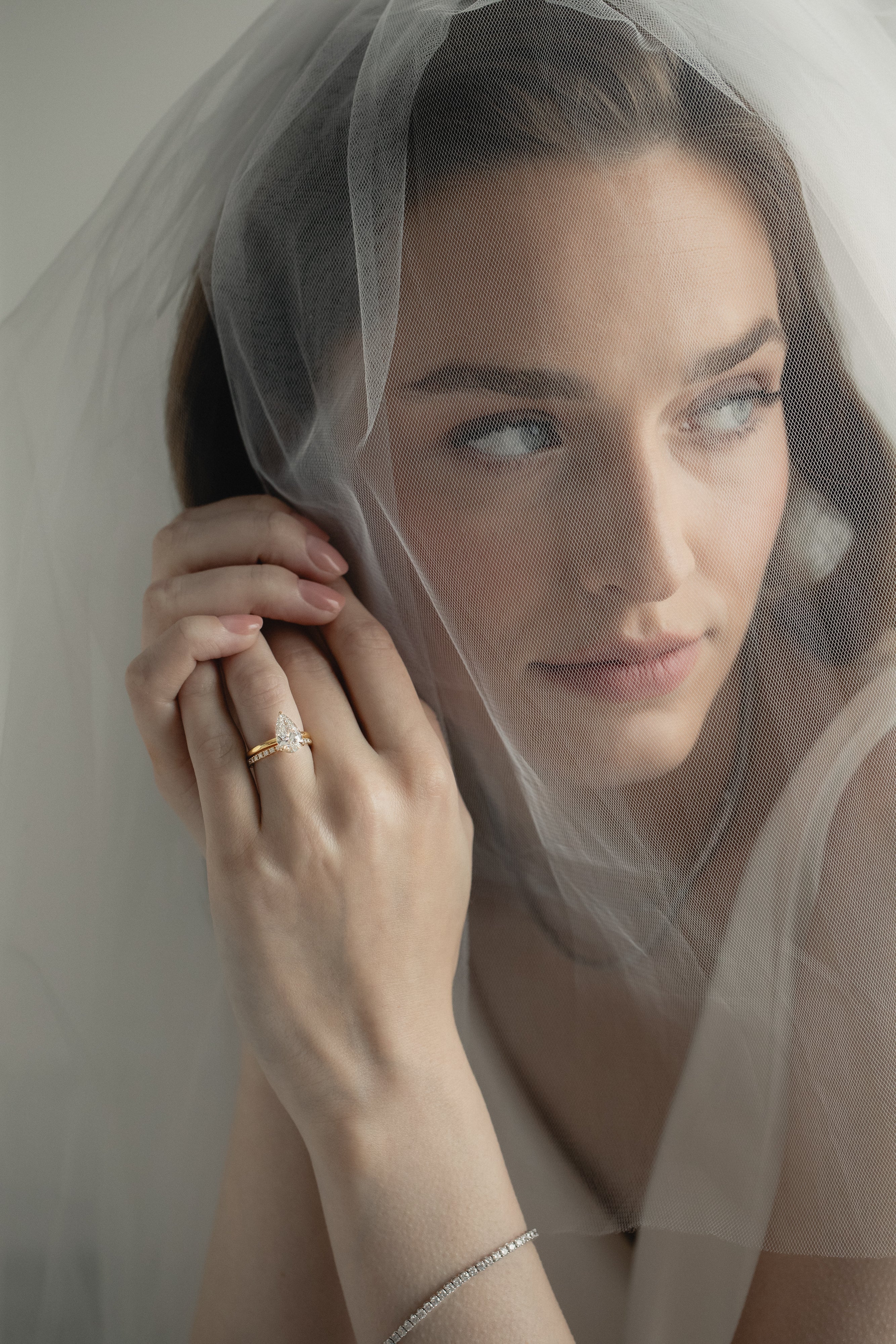 An editorial portrait of a bride looking through a sheer wedding veil, elegantly showcasing a KARAAT pear-shaped diamond solitaire engagement ring stacked with a matching white gold wedding band.