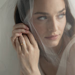 An editorial portrait of a bride looking through a sheer wedding veil, elegantly showcasing a KARAAT pear-shaped diamond solitaire engagement ring stacked with a matching yellow gold wedding band.