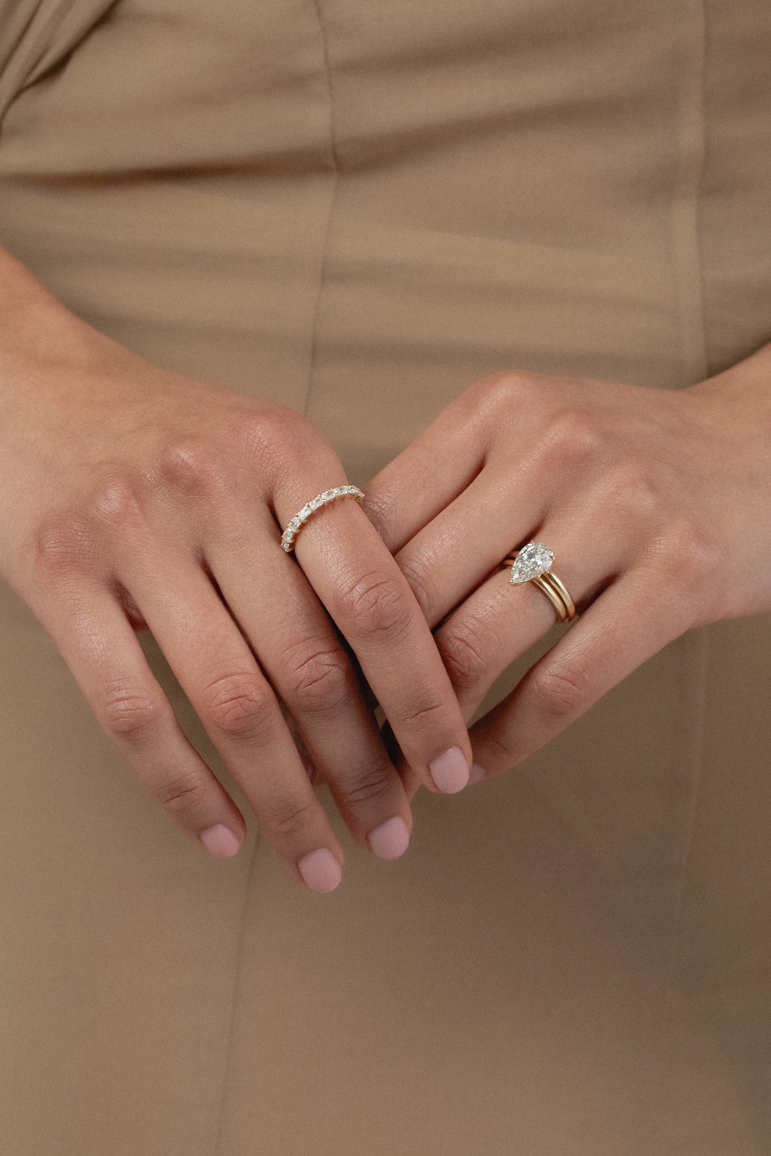 A close-up of a model's hands crossed against a beige dress, styled with a white gold KARAAT pear-shaped diamond solitaire ring stacked with a gold band on one hand, and a radiant-cut diamond half eternity band on the other.