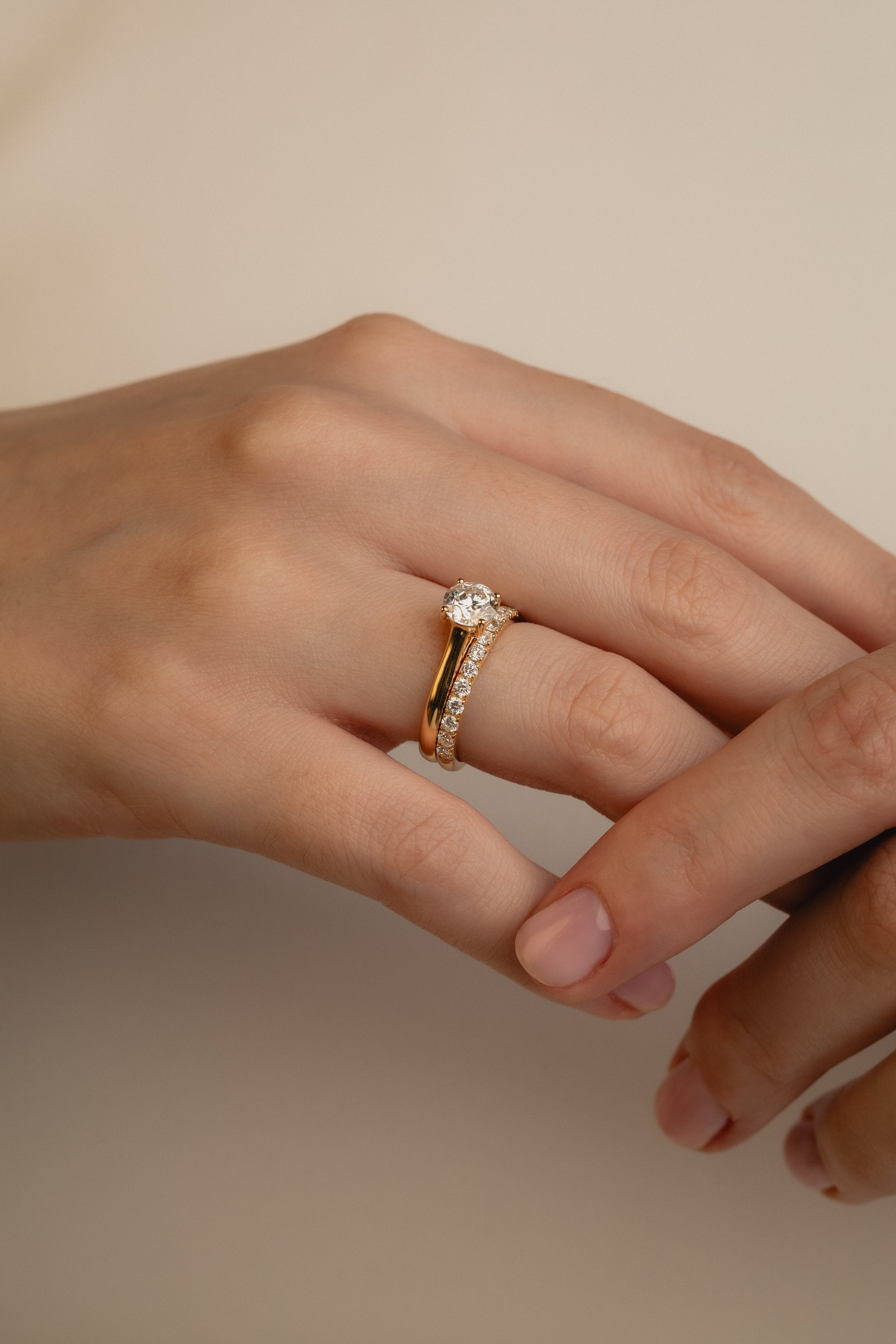 A soft-focus luxury bridal shot of a model's hand displaying a brilliant round solitaire diamond engagement ring nested flush beside the statement Pavé Diamond Ring III on a classic yellow gold band.