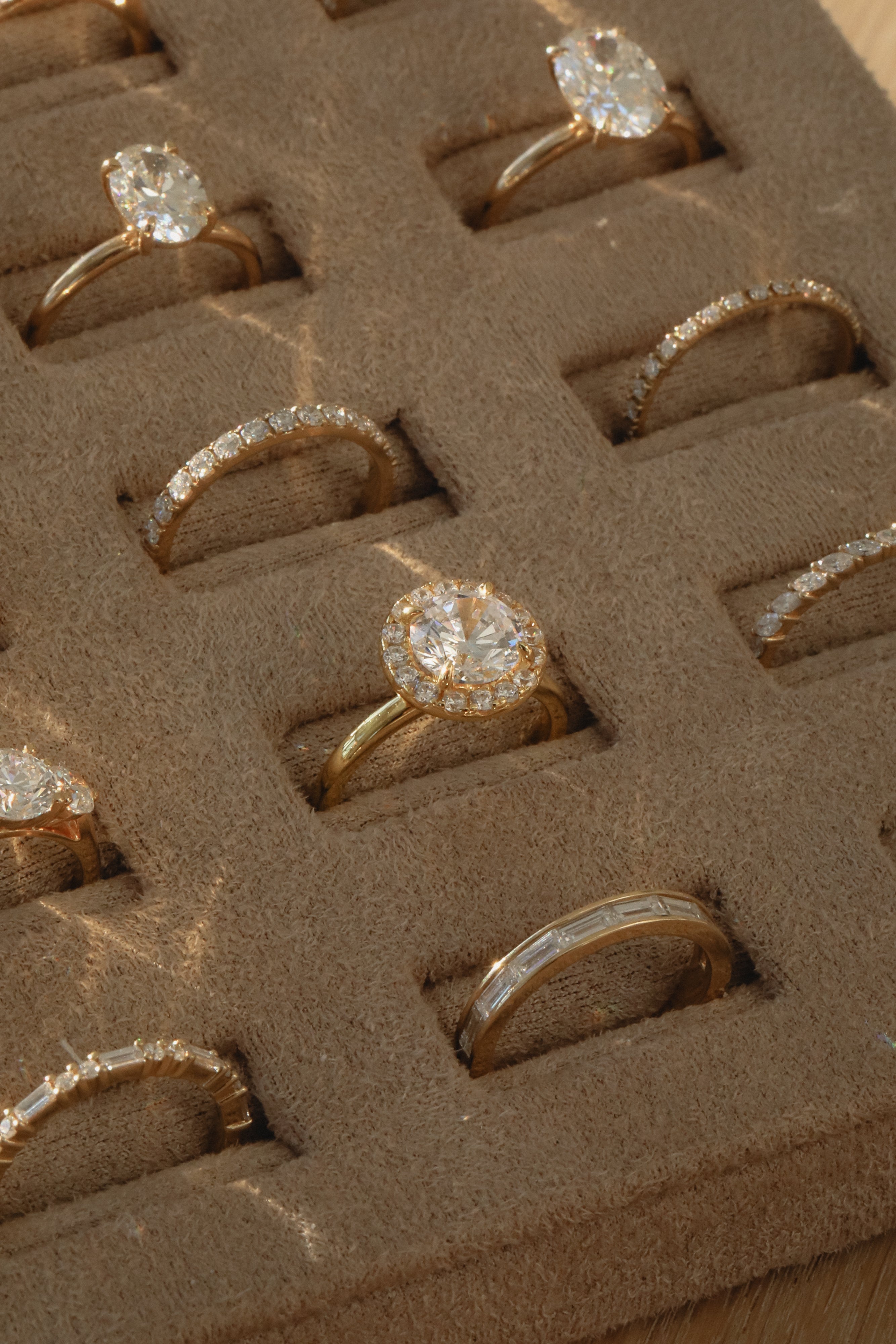A close-up view of a KARAAT 18k white gold round solitaire diamond engagement ring with a pavé halo, beautifully displayed inside a plush beige velvet jewelry organizer tray alongside other wedding and engagement rings.