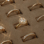 A close-up view of a KARAAT 18k rose gold round solitaire diamond engagement ring with a pavé halo, beautifully displayed inside a plush beige velvet jewelry organizer tray alongside other wedding and engagement rings.