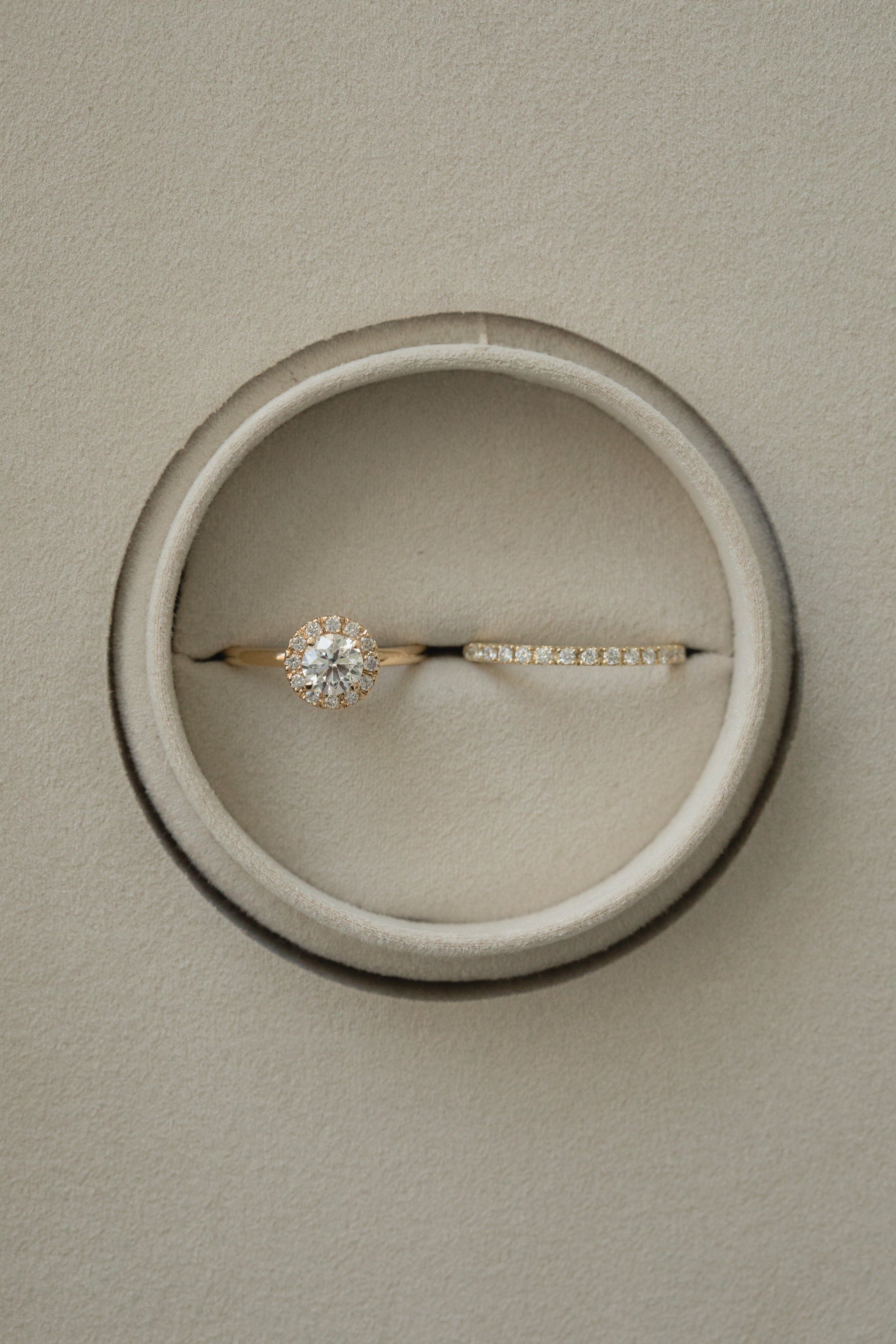 A flat-lay, top-down photograph of a rose gold KARAAT round solitaire halo diamond engagement ring paired with a matching sparkling diamond wedding band, perfectly nestled side-by-side inside a round taupe velvet ring box.