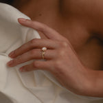 A close-up view of a model’s hand resting against a cream silk blouse, highlighting the elegant side profile, raised setting, and classic 18k gold band of the rose gold KARAAT round solitaire halo diamond ring.