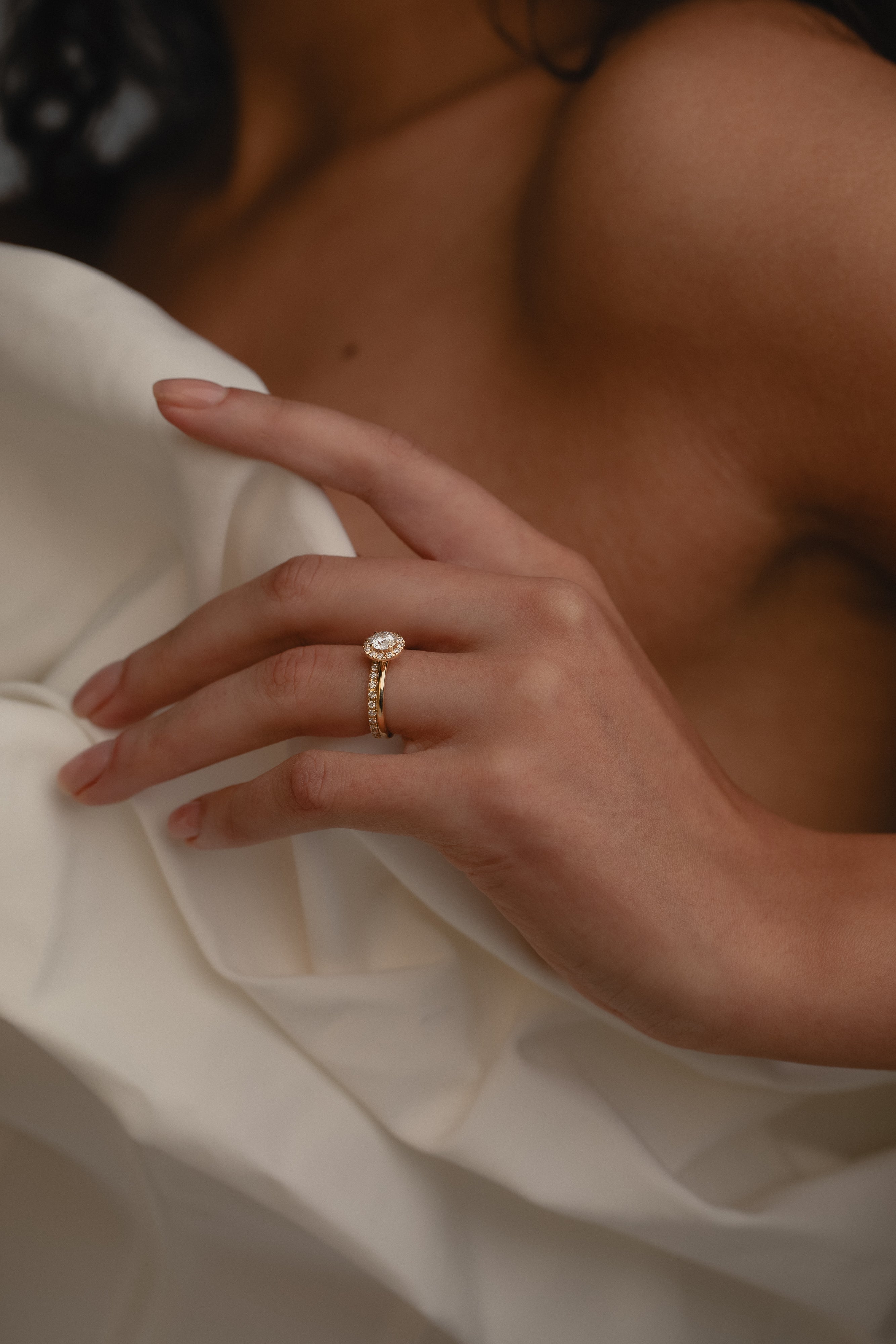 A close-up view of a model’s hand resting against a cream silk blouse, highlighting the elegant side profile, raised setting, and classic 18k gold band of the rose gold KARAAT round solitaire halo diamond ring.