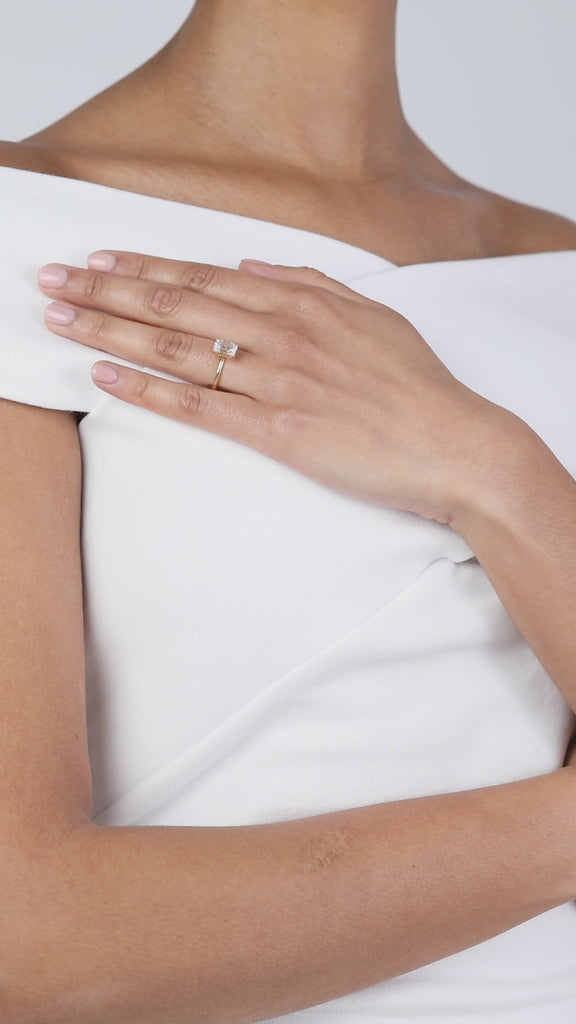 A elegant product video showcasing a KARAAT emerald-cut solitaire diamond engagement ring on an 18k yellow gold band. The model wears a white off-the-shoulder top and rests her hand on her chest, beautifully capturing the clean lines and flash of the diamond.