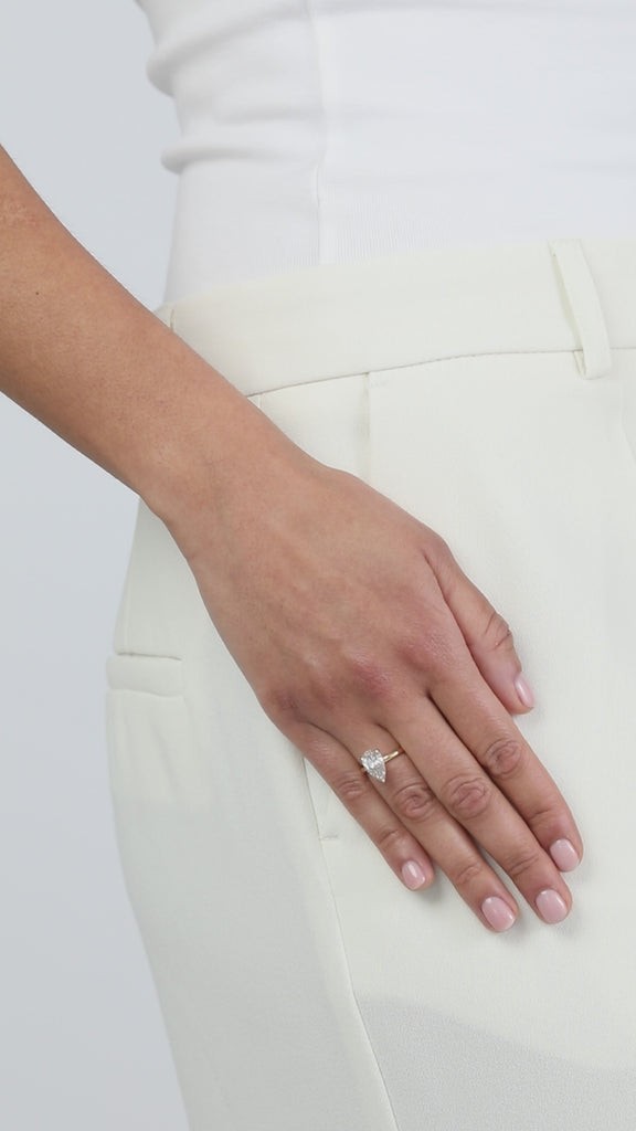 A clean product video showcasing a KARAAT pear-shaped diamond solitaire engagement ring on an 18k rose gold band. The model has her hand casually placed in the pocket of tailored white trousers, capturing the brilliant sparkle and modern elegance of the ring against the crisp white fabric.