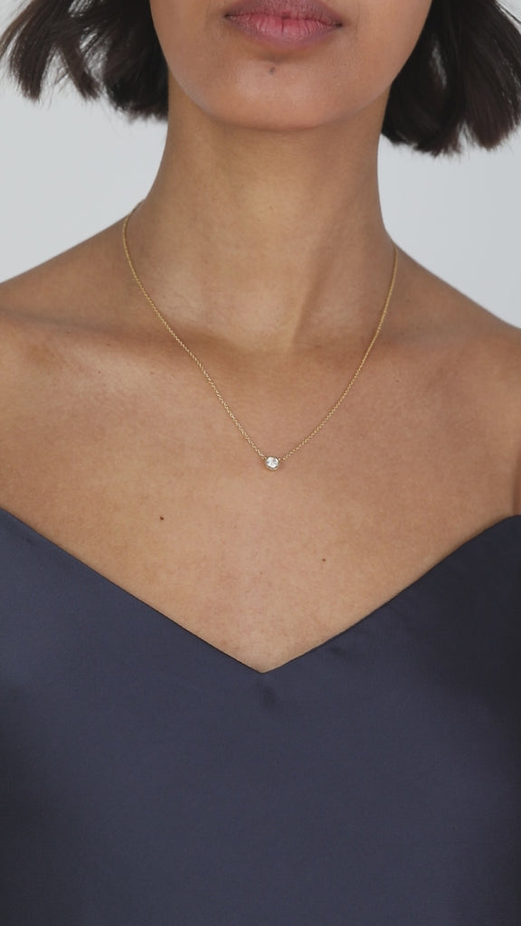 A lifestyle video displaying the delicate KARAAT diamond bezel solitaire necklace on a model's neckline, showcasing how the sparkling round-cut diamond catches the light against a fine rose gold chain.